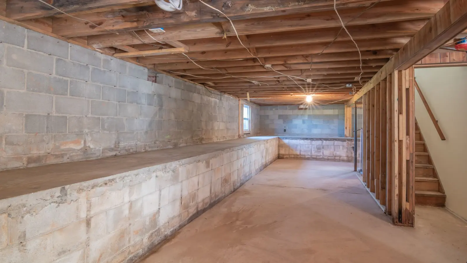 Unfinished basement with concrete block walls, open wood floor joists and a stairway