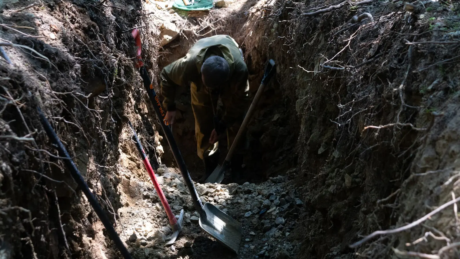 Worker digging a deep, narrow trench with shovels resting against the sides